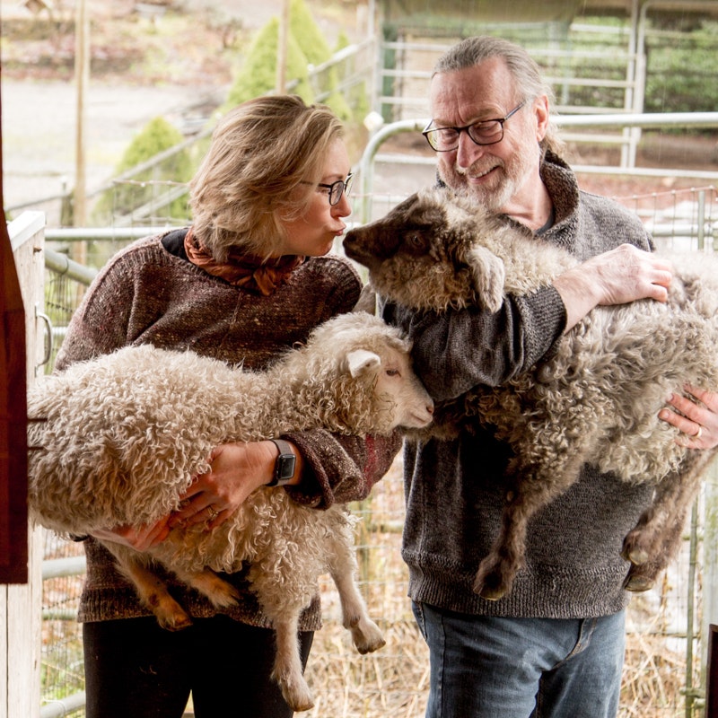 Woman in brown sweater, man in brown sweater, both hold curly goats, woman kisses the goat in the man's arms