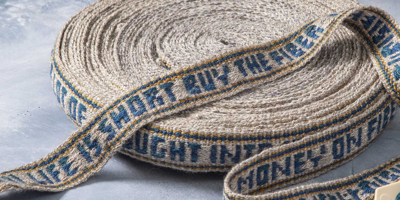 A beige handwoven band with blue text rests on a blue-gray background. The text woven into the band reads, "Life is short buy the fiber."
