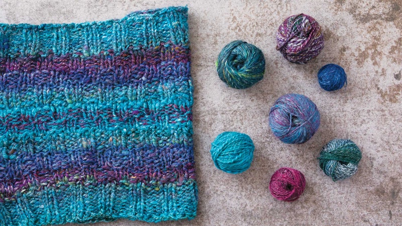 Turquoise, purple, and magenta cowl with small balls of yarn sitting next to it on a beige background.