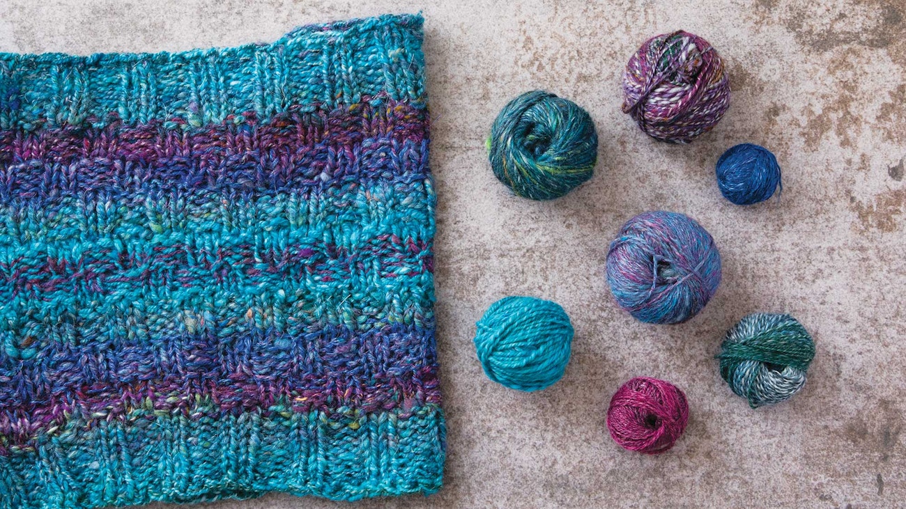 Turquoise, purple, and magenta cowl with small balls of yarn sitting next to it on a beige background.