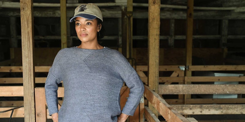 A woman stands in a dark barn wearing a blue-gray sweater with a zig-zag detail down the front. She is wearing a baseball cap and looks into the distance.