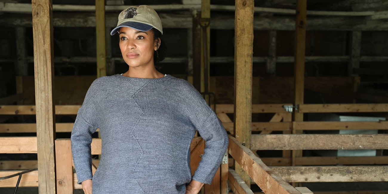 A woman stands in a dark barn wearing a blue-gray sweater with a zig-zag detail down the front. She is wearing a baseball cap and looks into the distance.