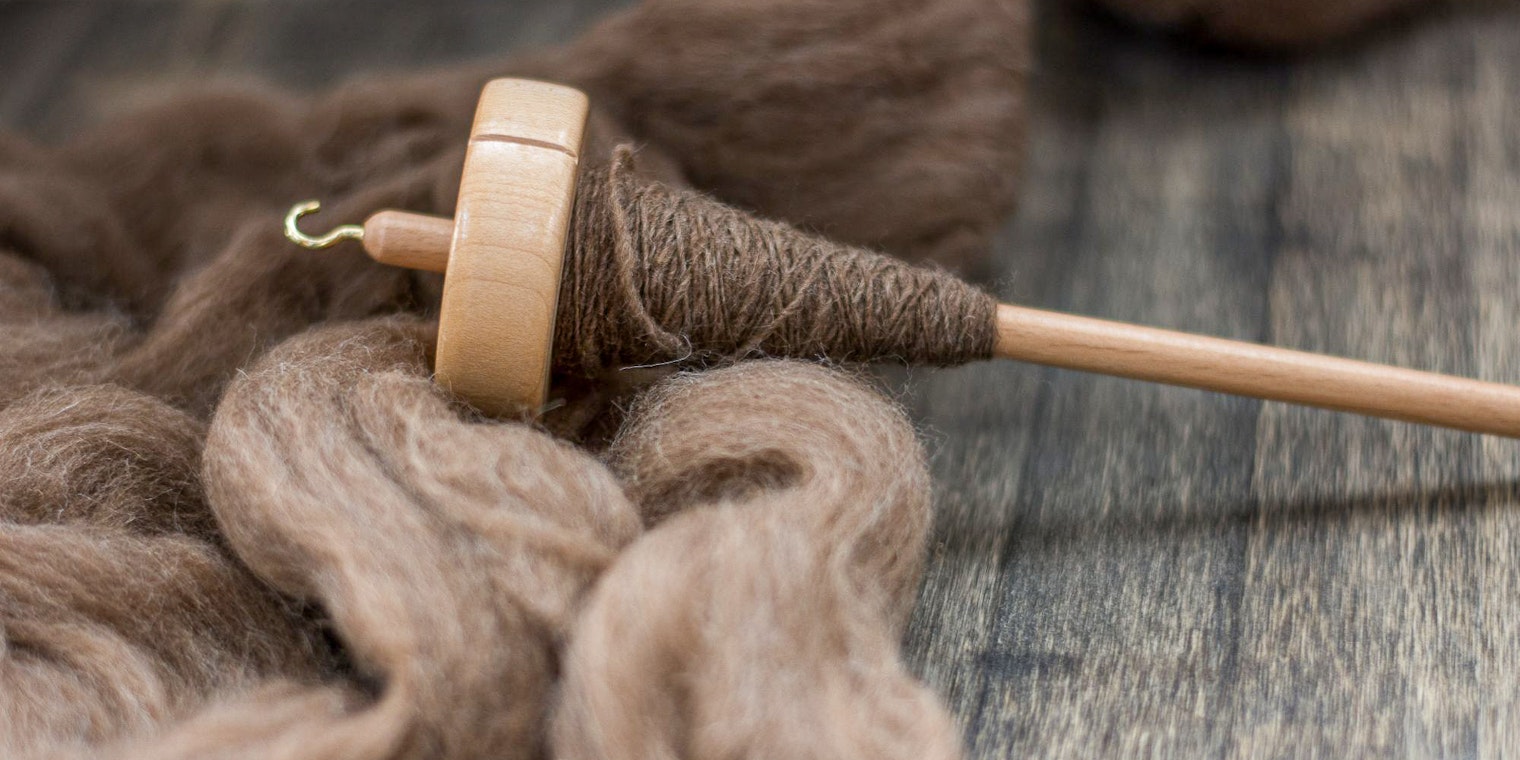 A spindle with spun fiber rests on top of natural brown unspun wool.