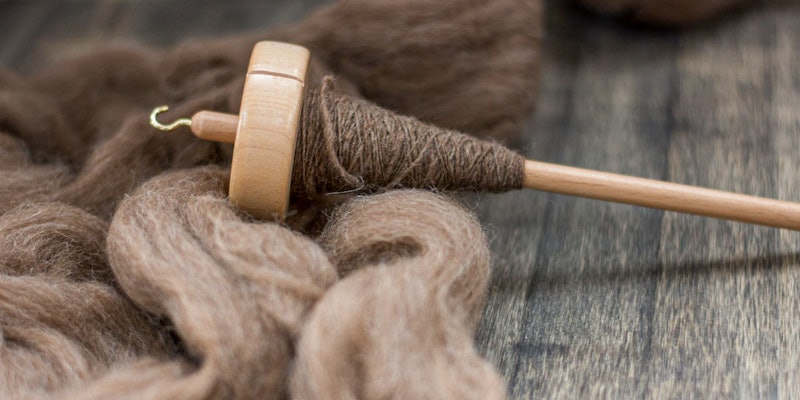 A spindle with spun fiber rests on top of natural brown unspun wool.