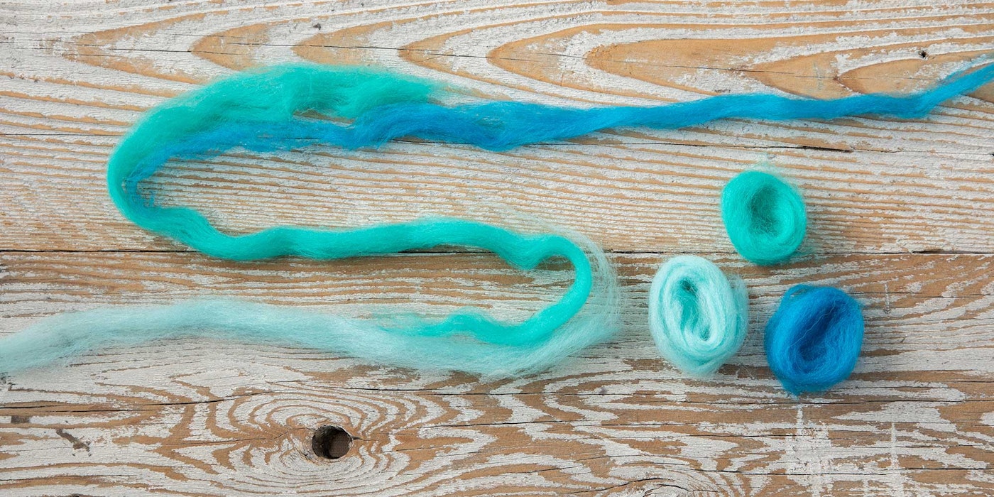 Thin ribbons of roving with a smooth color change from pale to dark turquoise blue rest on a distressed wood background.