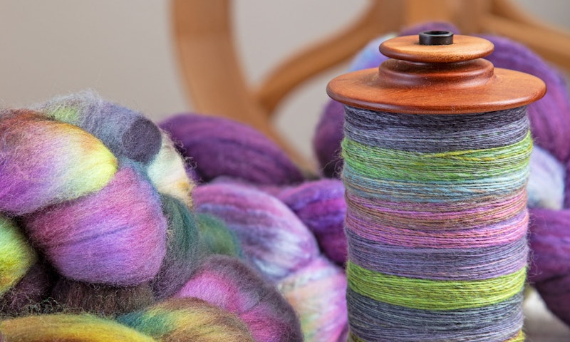 A closeup of fiber and handspun yarn. A wheel is out of focus in the background