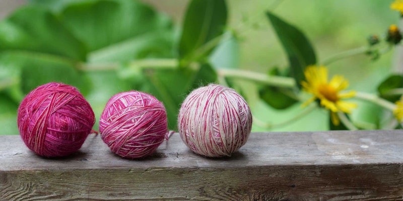 Three plying balls, in varying saturations of magenta, rest on a garden rail with yellow flowers in the background.