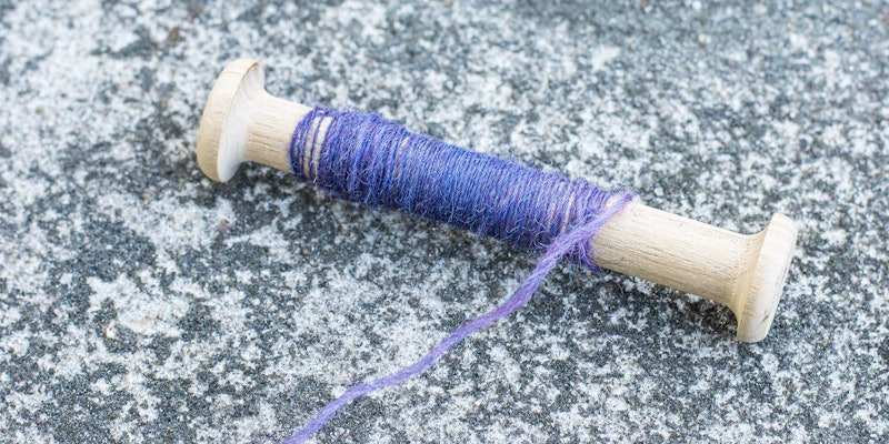 A wooden bobbin with purple yarn rests on a concrete surface.
