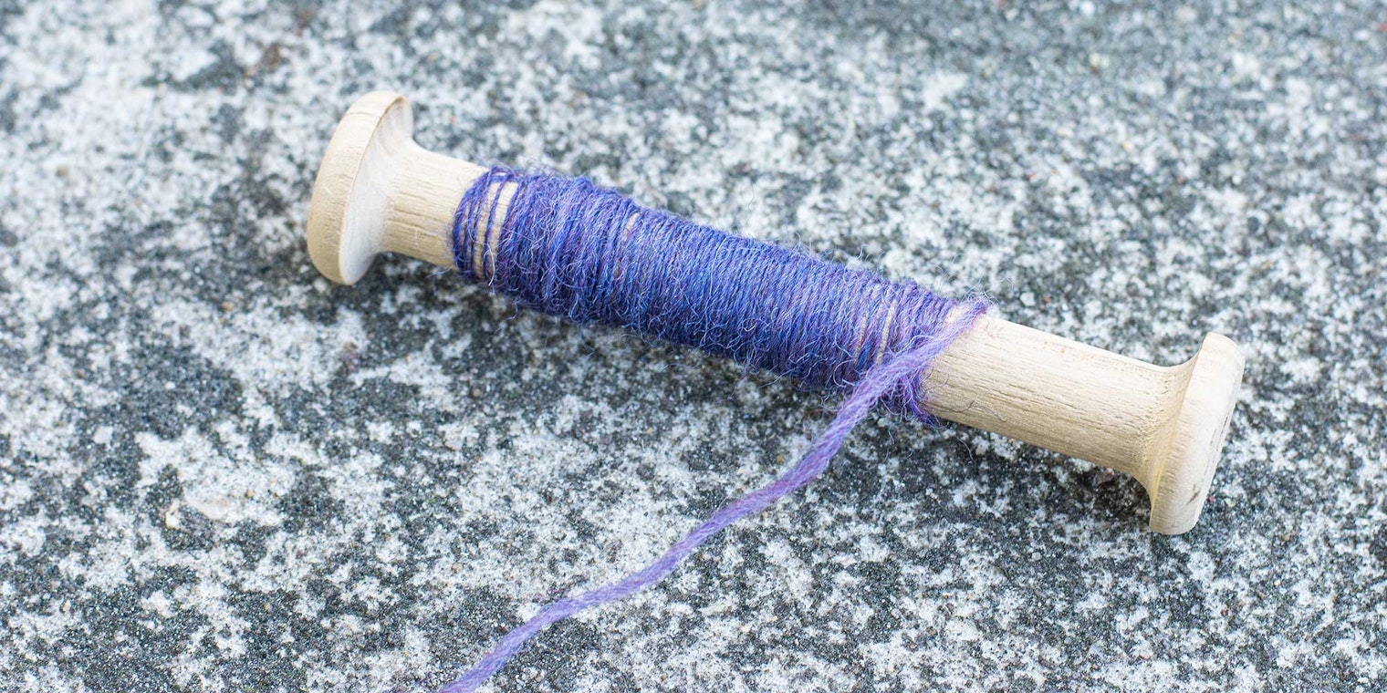 A wooden bobbin with purple yarn rests on a concrete surface.