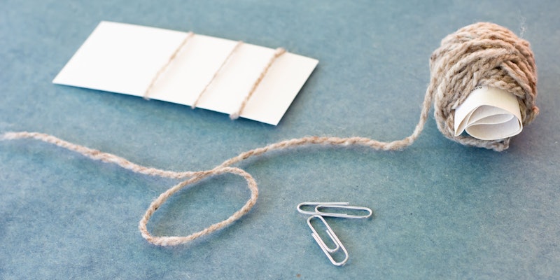 Two paperclips, a ball of yarn, and a card with deconstructed yarn on a blue surface