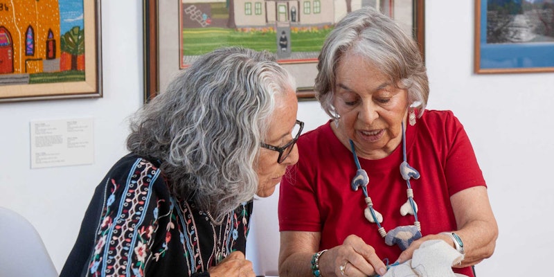 A woman in a red shirt is teaching embroidery techniques to a woman in a black patterned shirt.