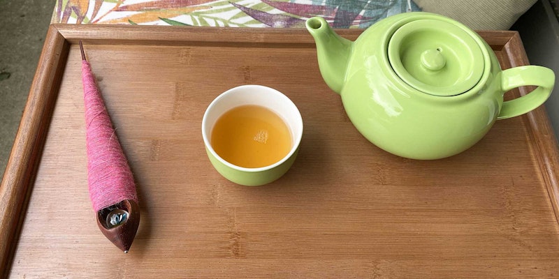 A supported spindle with pink yarn rests on a tray next to a cup of tea and a lime green teapot.