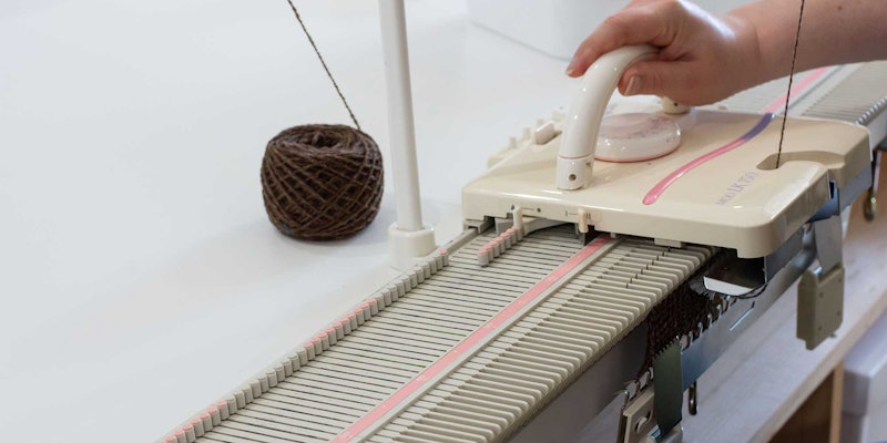 A hand is pushing a carriage across a flatbed knitting machine. The machine is threaded with brown handspun yarn.