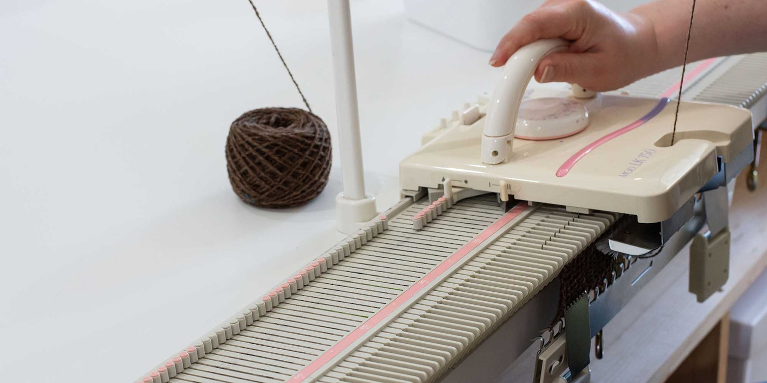 A hand is pushing a carriage across a flatbed knitting machine. The machine is threaded with brown handspun yarn.