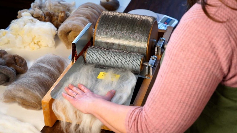A hand feeds fiber onto a drumcarder