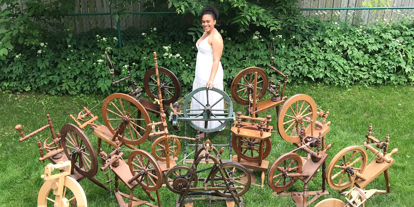 A woman wearing a white dress stands in the garden. She is surrounded by 15 spinning wheels.