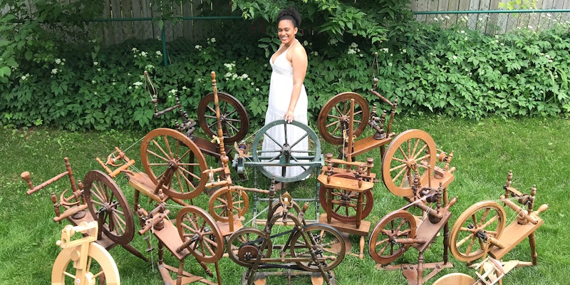A woman wearing a white dress stands in the garden. She is surrounded by 15 spinning wheels.