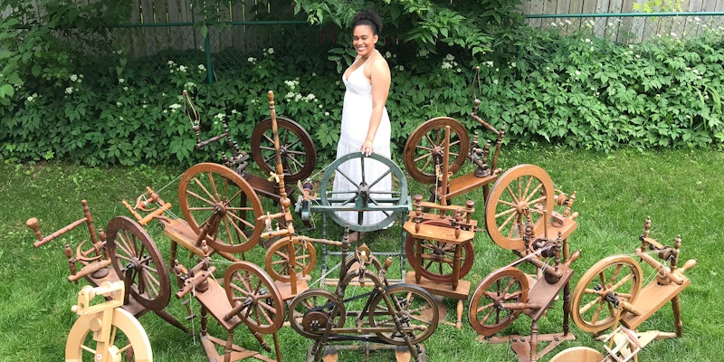 A woman wearing a white dress stands in the garden. She is surrounded by 15 spinning wheels.