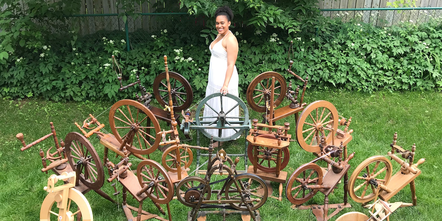A woman wearing a white dress stands in the garden. She is surrounded by 15 spinning wheels.