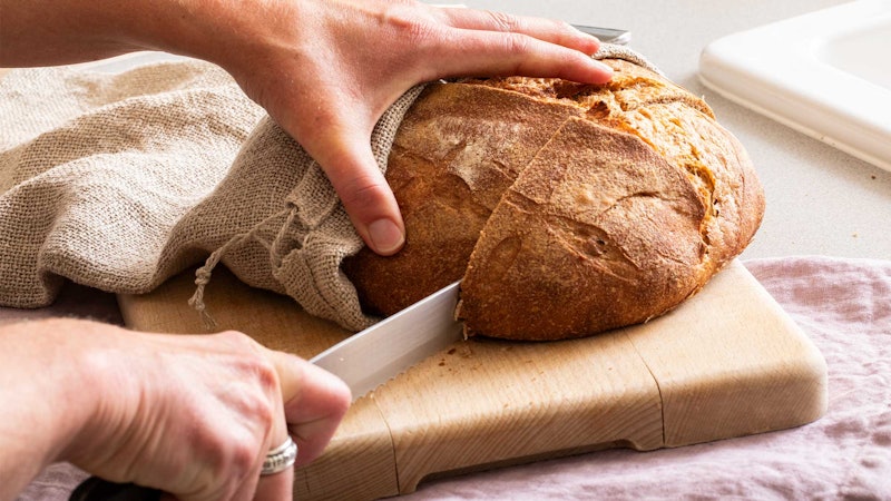 A pair of hands cutting sourdough bread on a wooden cutting board.