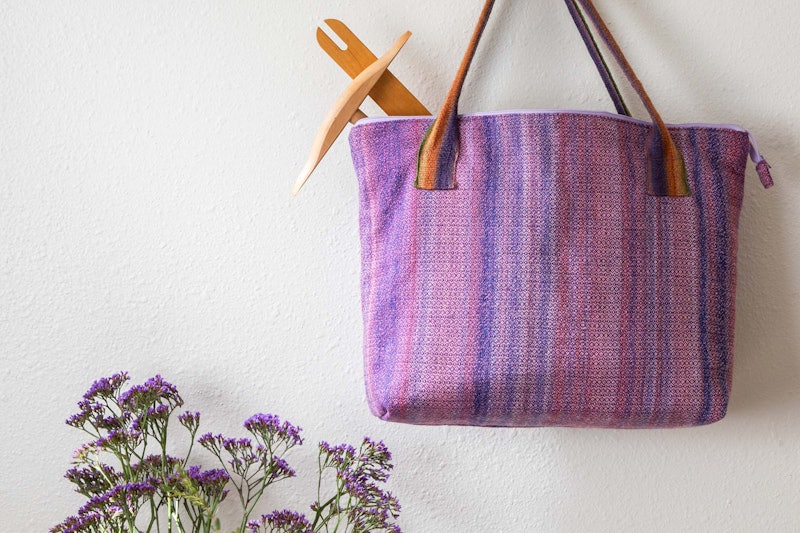 A striped tote bag with a niddy noddy and weaving shuttle poking out of it. There are purple flowers in the bottom left-hand corner of the photo.