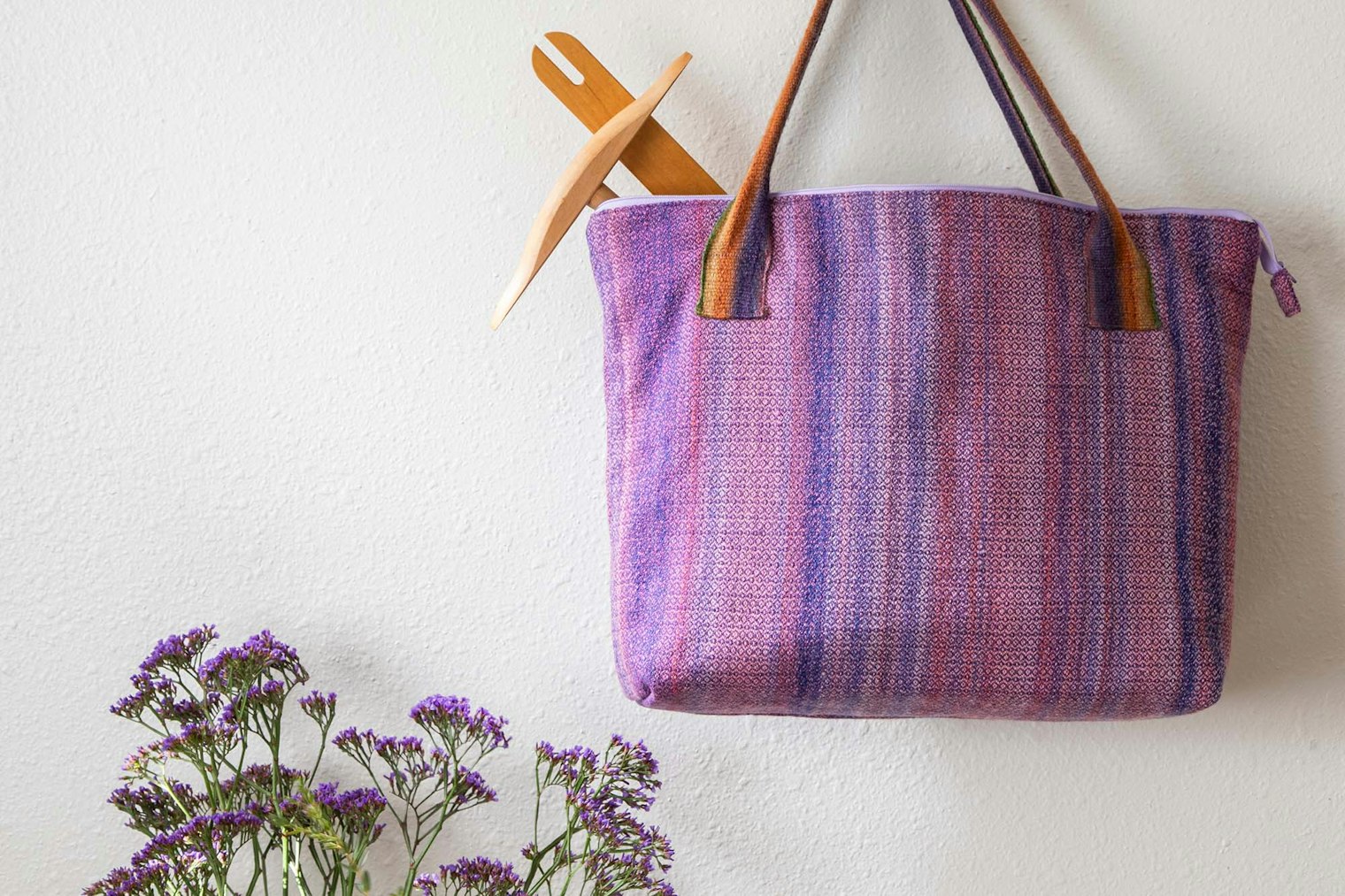 A striped tote bag with a niddy noddy and weaving shuttle poking out of it. There are purple flowers in the bottom left-hand corner of the photo.