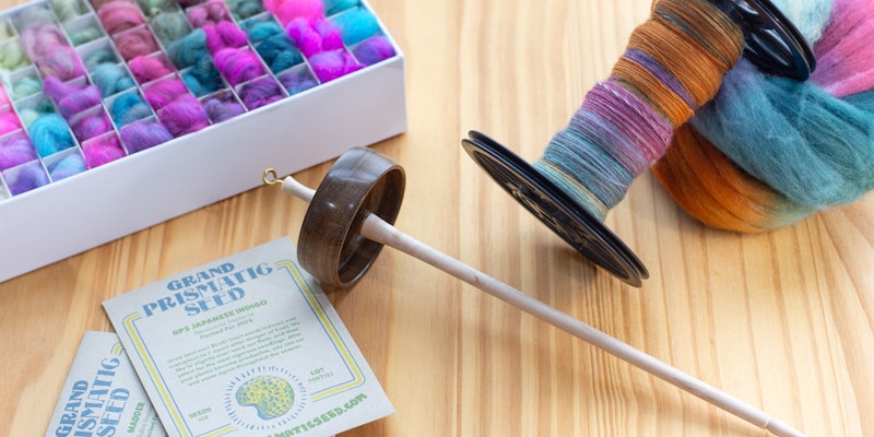 A colorful array of spinning fiber, spun fiber on a bobbin, a spindle, and seed packets on a wooden surface.