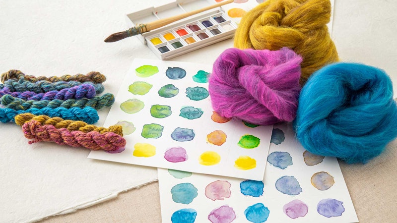 Watercolor samples, fiber and yarn. There is a paintbrush and pan of watercolors in the background.