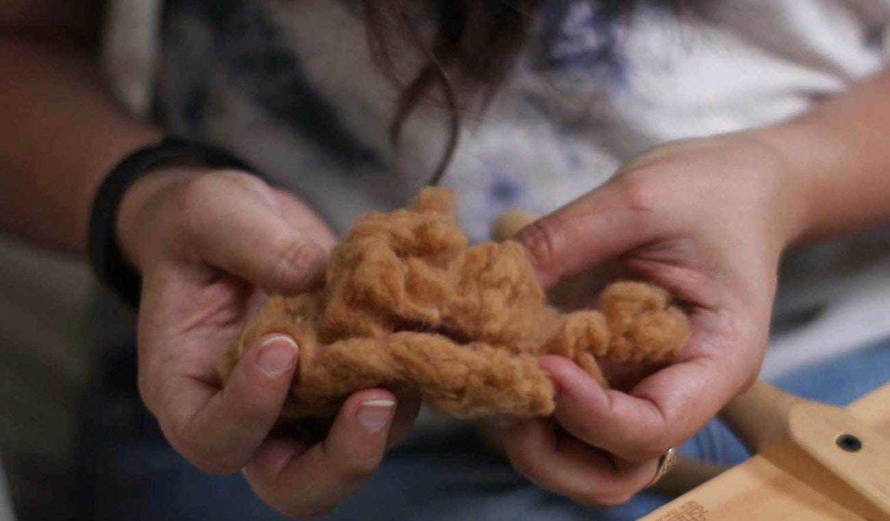 Hands holding brown cotton. A pair of handcarders can be seen in the lower right edge of the photo.