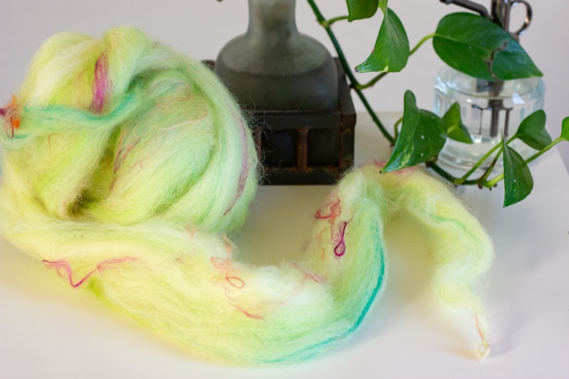 A lime green ball of roving with pink and turquoise accents is shown on a white surface. A plant and mister are in the background.