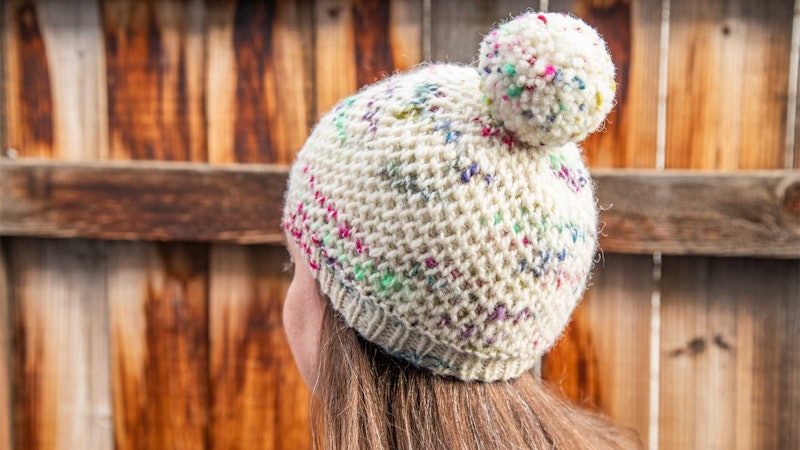 The back of a person's head. The person is wearing a speckled beanie with a pom pom.