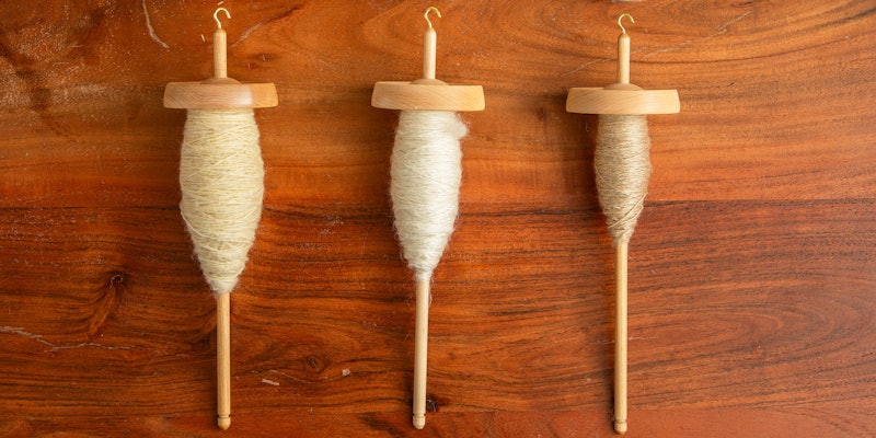 Three drop spindles on a wood surface with different amounts of spun yarn on each of them.