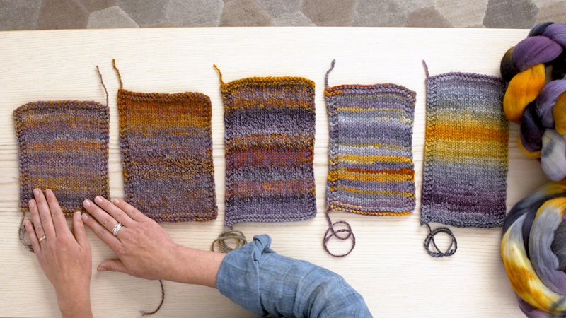 Five knitted swatches on a table with spinning fiber peeking in on the right. Hands are touching the leftmost knitted swatch.
