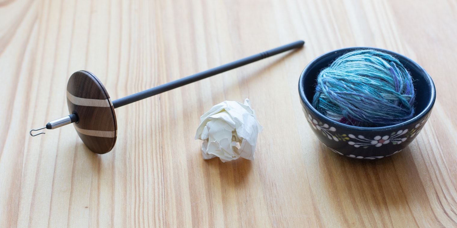 A spindle, wadded up piece of paper, and a ball of yarn in a ceramic bowl sit on a wooden surface.