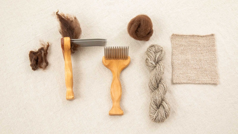 Wool and wool combs on a beige surface. On the right is a skein of yarn and a knitted swatch.