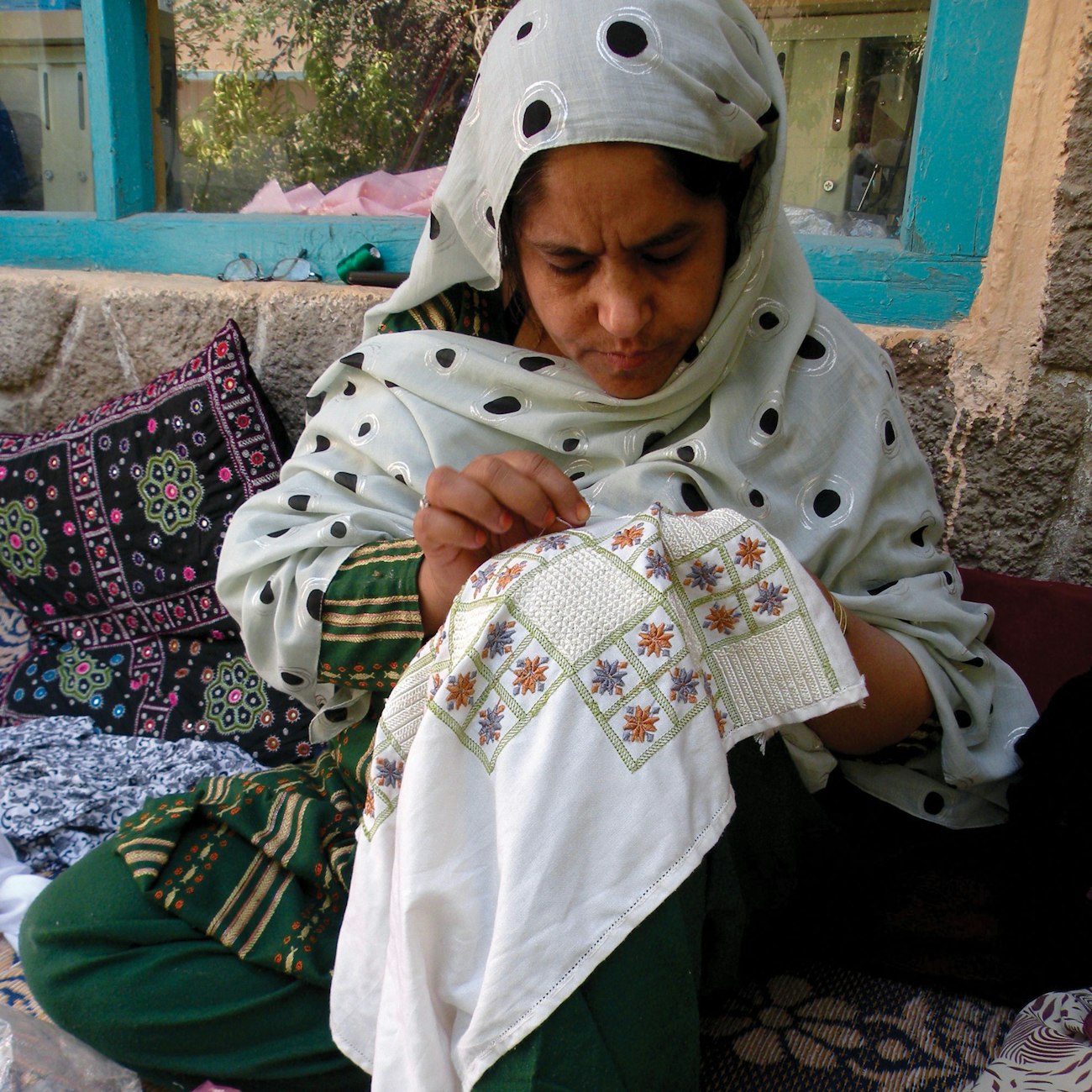 A woman with a gray spotted headscarf, seated on the ground, embroiders on a white ground cloth.