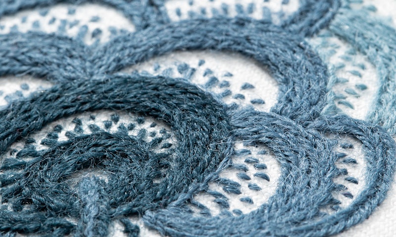 Detail of wool crewelwork embroidery, in blue thread on white fabric.