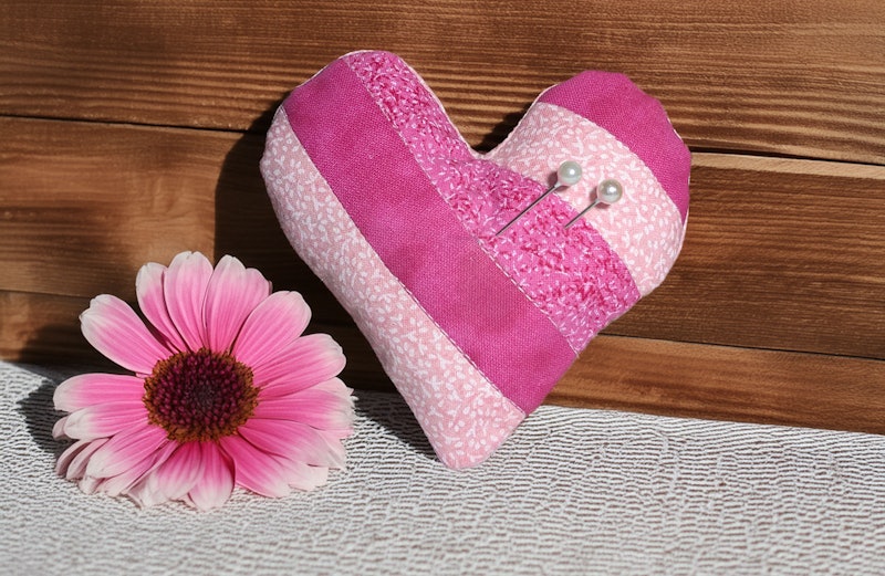 A quilted heart with two pins it it, propped up on a table and sitting next next to a pink flower.