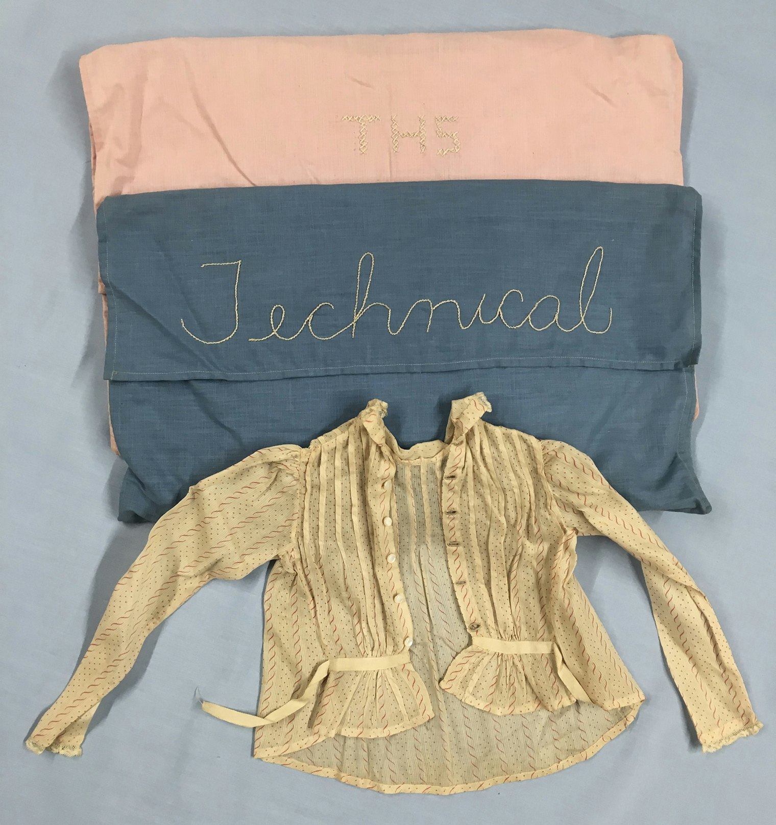 Pierce student sample. Each of Miss Pierce’s students sewed a bag in which they kept their samples. The shirt is a small-scale model of a shirtwaist, worn around 1900. The back, complete with handsewn buttonholes, faces up in the photo; the longer front created the pigeon-breasted look popular at the time, with the belt sewn to the back tying around to the front. Donor: Lucy H. Pierce. Historic Textile and Costume Collection, University of Rhode Island, URI 2019.98.05a, b, c.