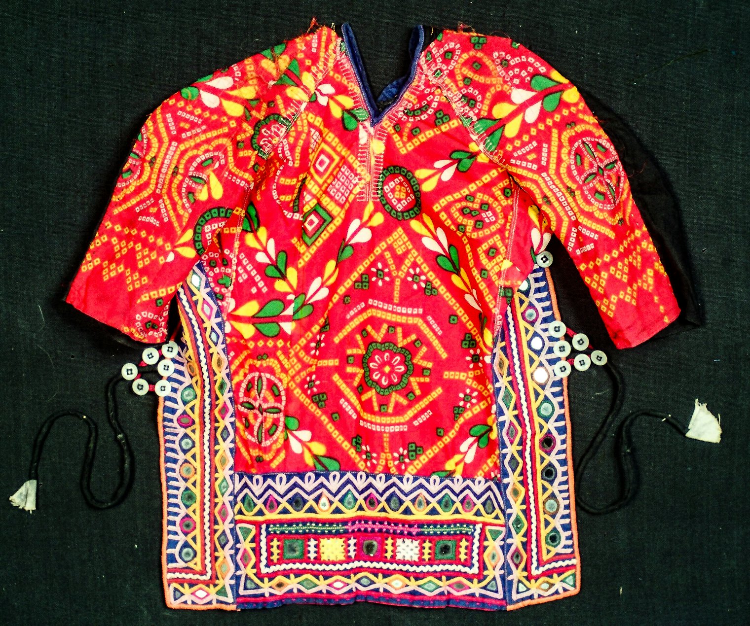 Kanchaliyu, unmarried girl’s everyday blouse,Viyar. Before girls married, their blouses did not have gathers at the breasts. Unmarried girls and young women wore blouses made of colorful and patterned fabric. The fabric of this blouse is synthetic with an industrial print derived from a bandhani resist-dyed pattern. By the 1960s and 70s, cheaper replicas began to replace traditionally used handmade fabrics. One of the first time-saving measures was substituting rickrack for makudiya—ants—traditionally represented by a white wavy chain-stitched pattern. That substitution dates this blouse to late 1970s to early 1980s. The sides and bottom border are embroidered with traditional Rabari patterns. Photo by Ketan Pomal, L.M. Studio, Bhuj, courtesy of Kala Raksha Museum.