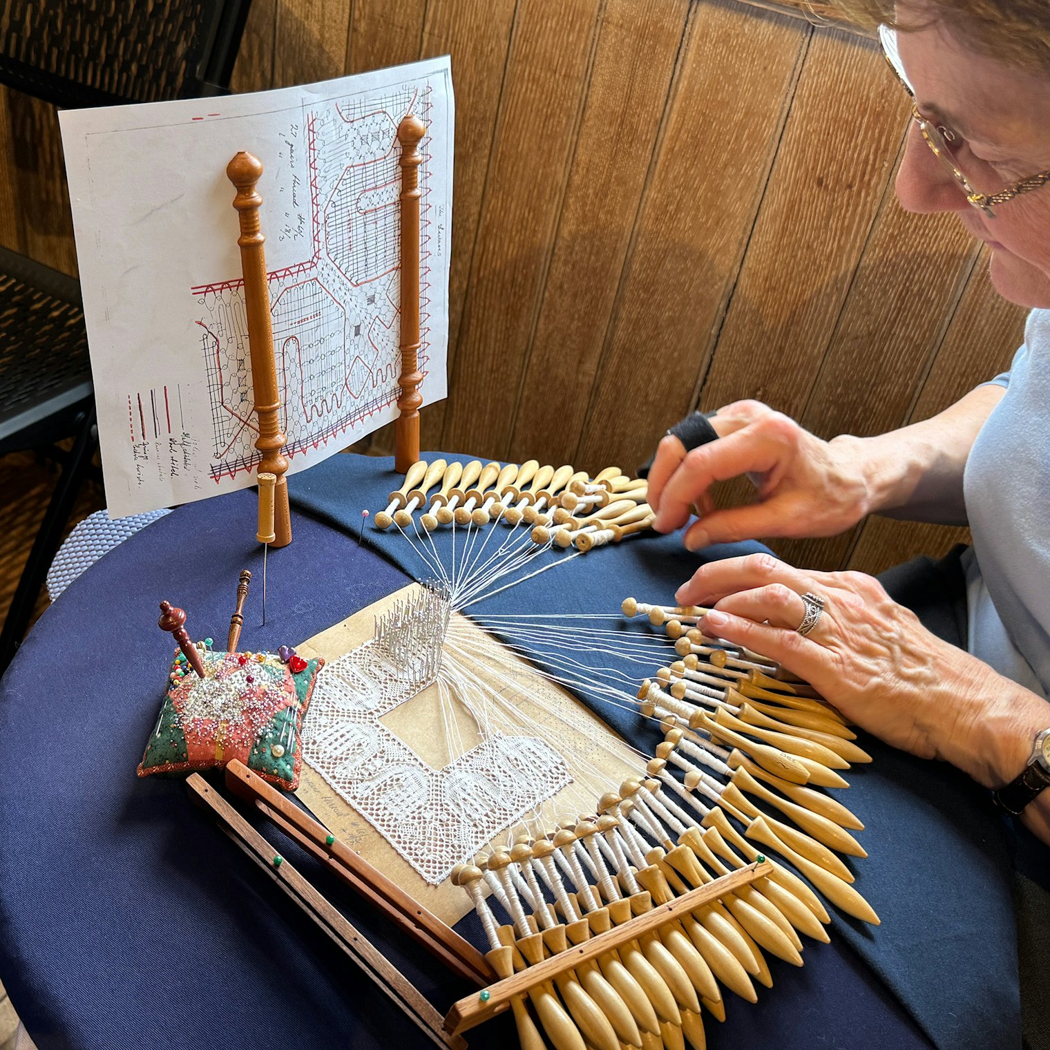 A lacemaker demonstrates her work in progress and her tools including the handy pattern holder to help keep thread paths in order.