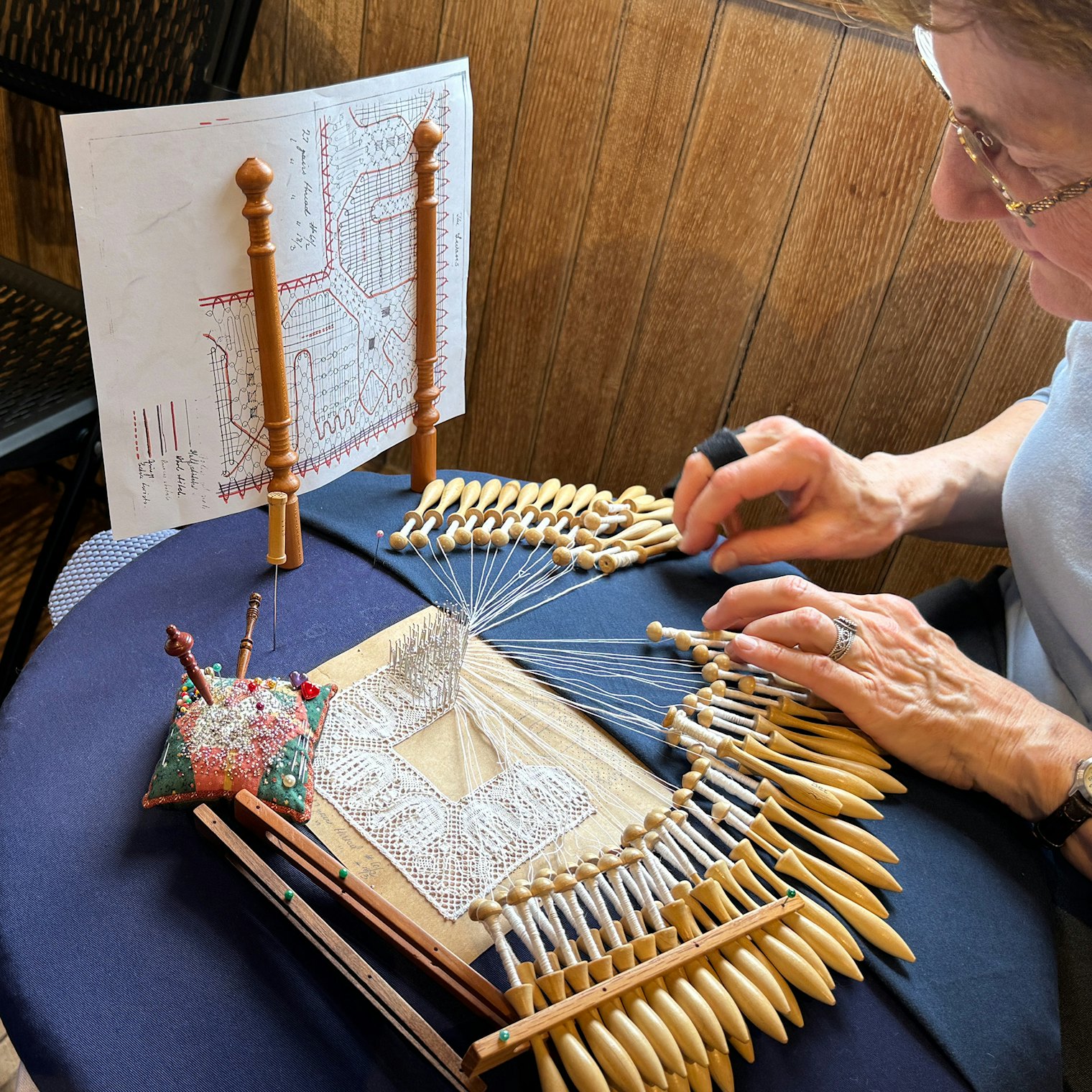 A lacemaker demonstrates her work in progress and her tools including the handy pattern holder to help keep thread paths in order.