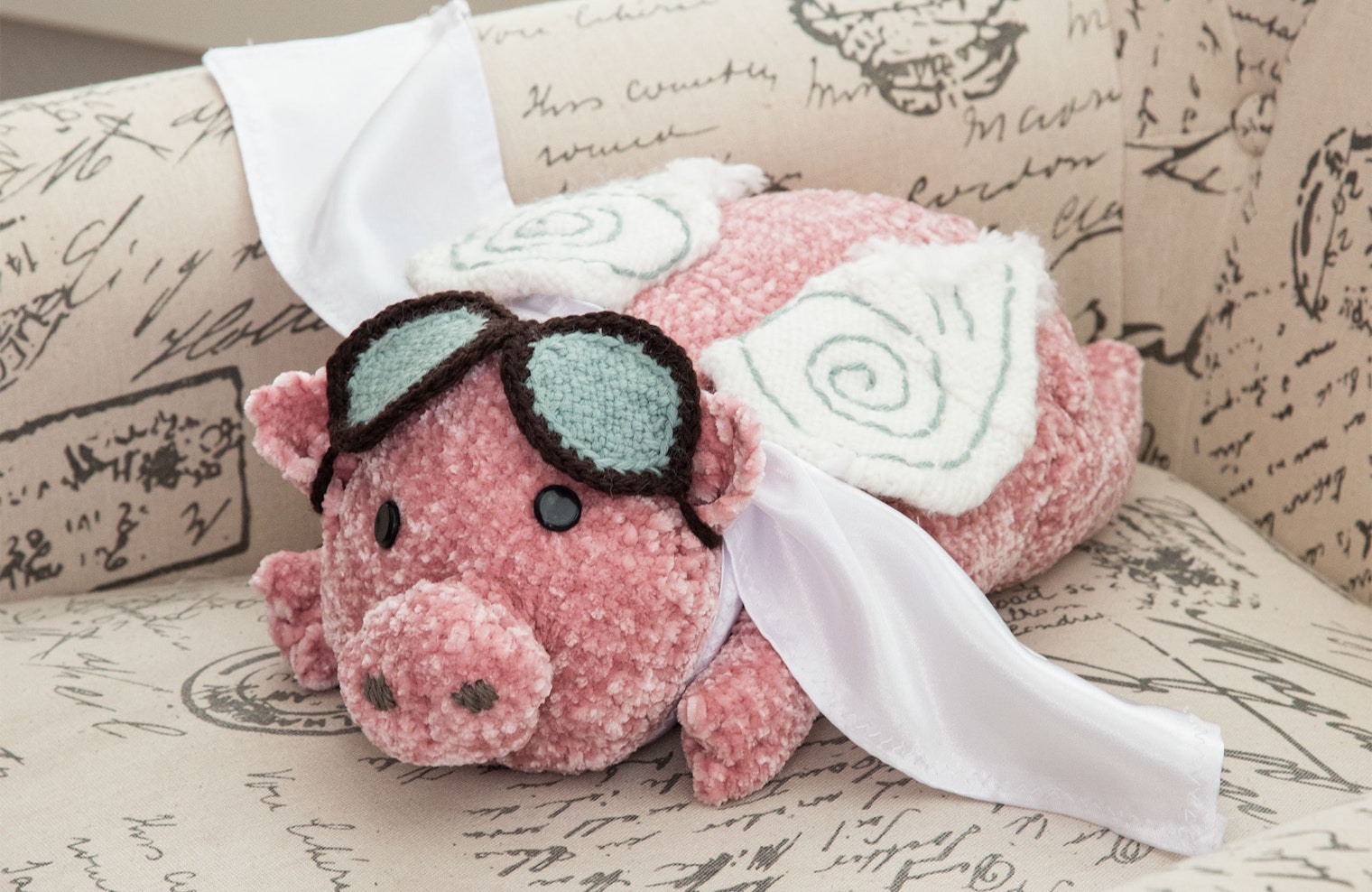 A pink stuffed pig toy on a chair, pin-loom-woven with wings, goggles, and cape