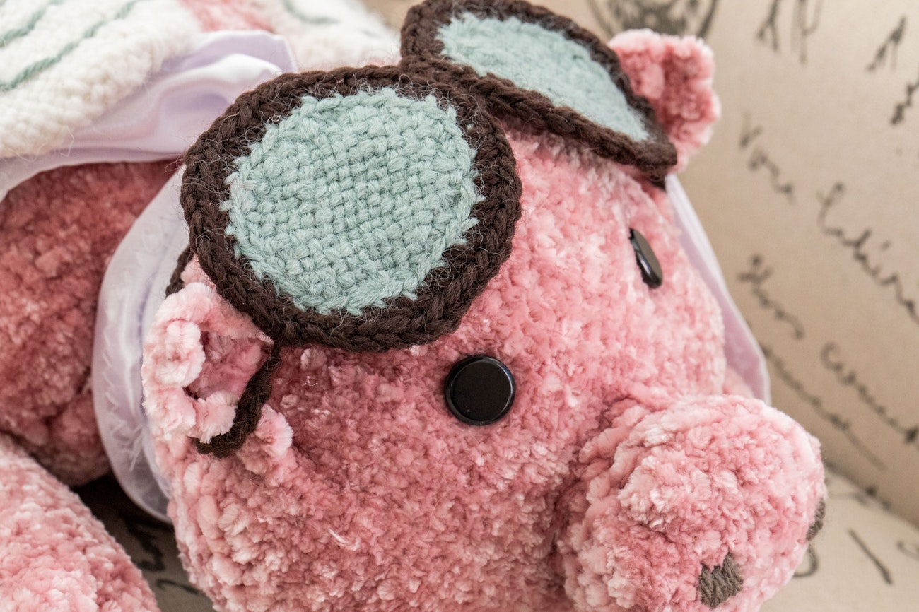 A pink stuffed pig toy, pin-loom-woven with wings, goggles, and cape