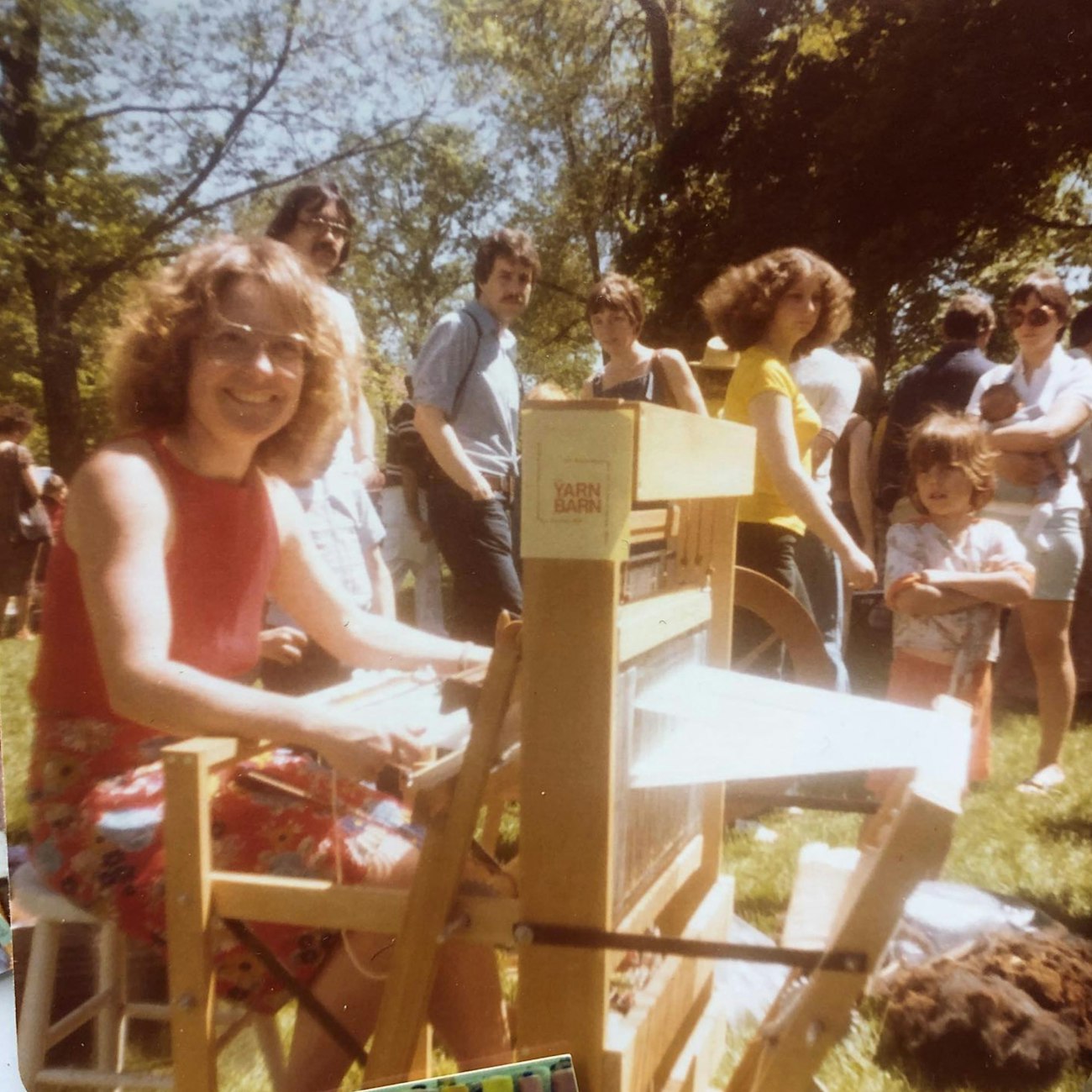 Woman in 1980s attire sitting at loom outside, smiling