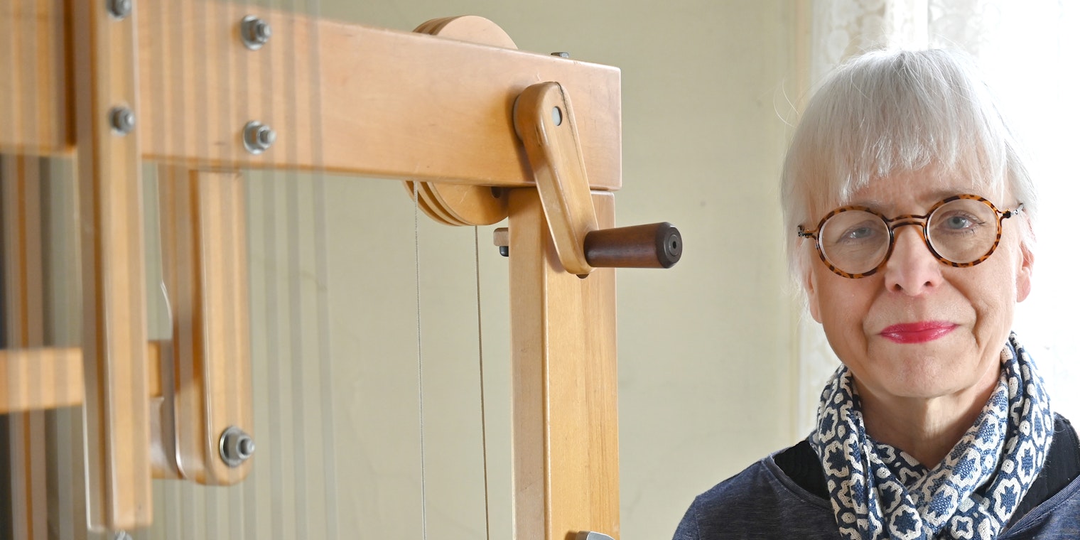 Woman wearing glasses stands next to a loom.