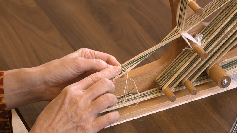 a weaver pointing to a partially woven inkle band with a tapestry needle weaving in the tail