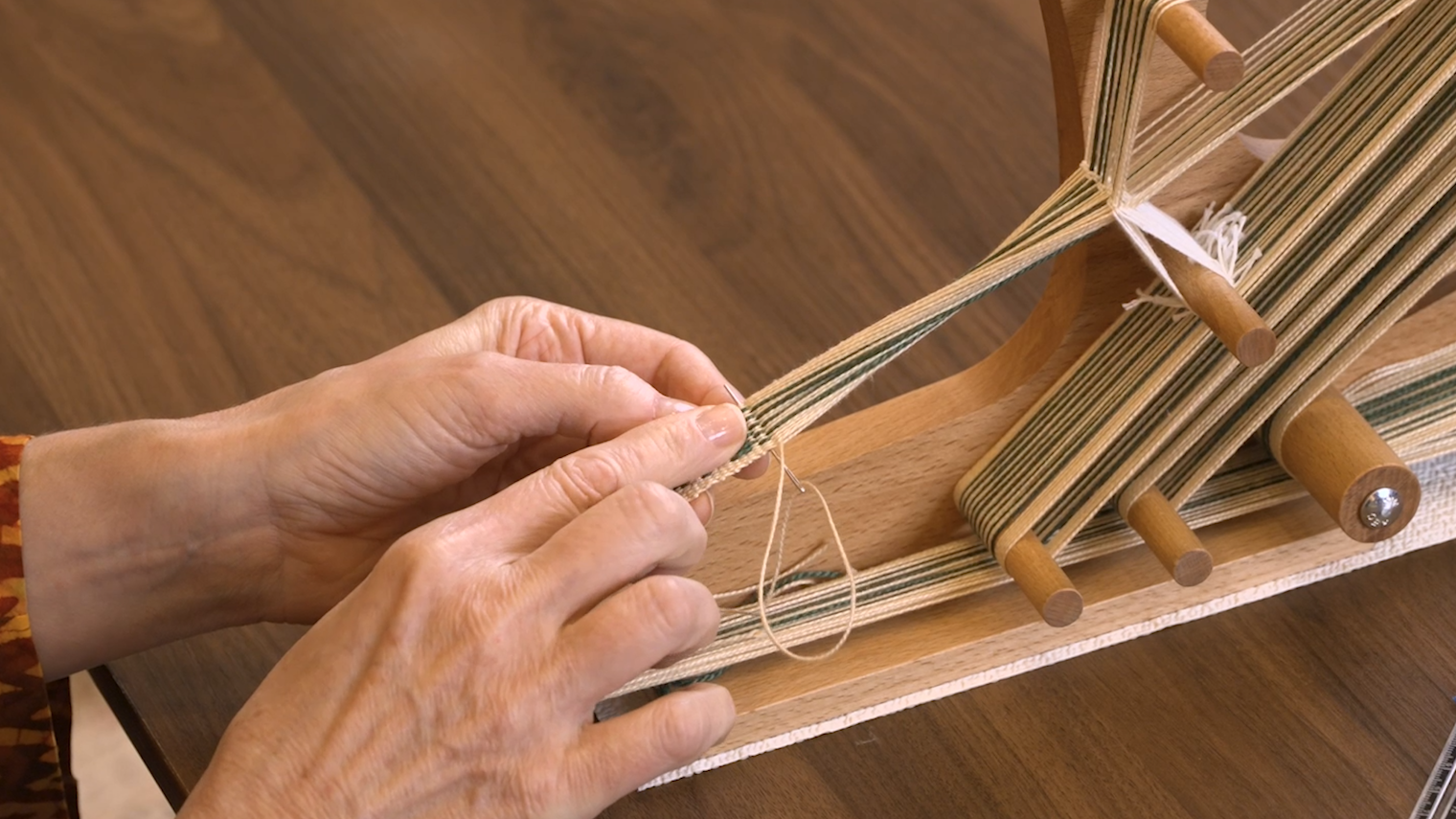 a weaver pointing to a partially woven inkle band with a tapestry needle weaving in the tail