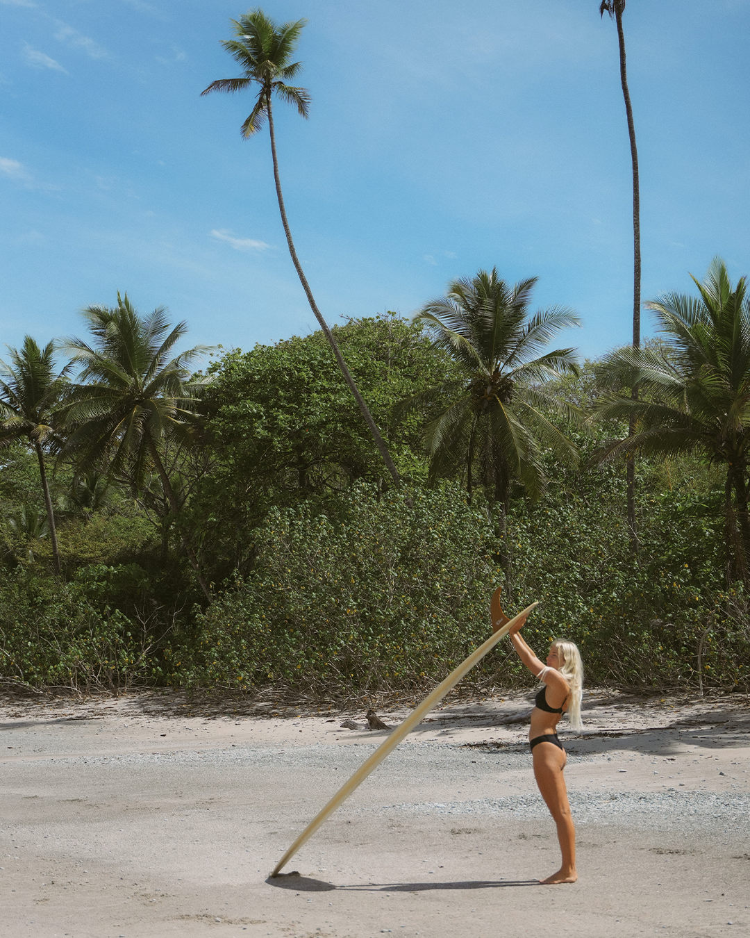 Gabriella wearing period-proof bikini bottoms with a matching bikini top, standing barefoot on a tropical beach, holding a surfboard under palm trees.