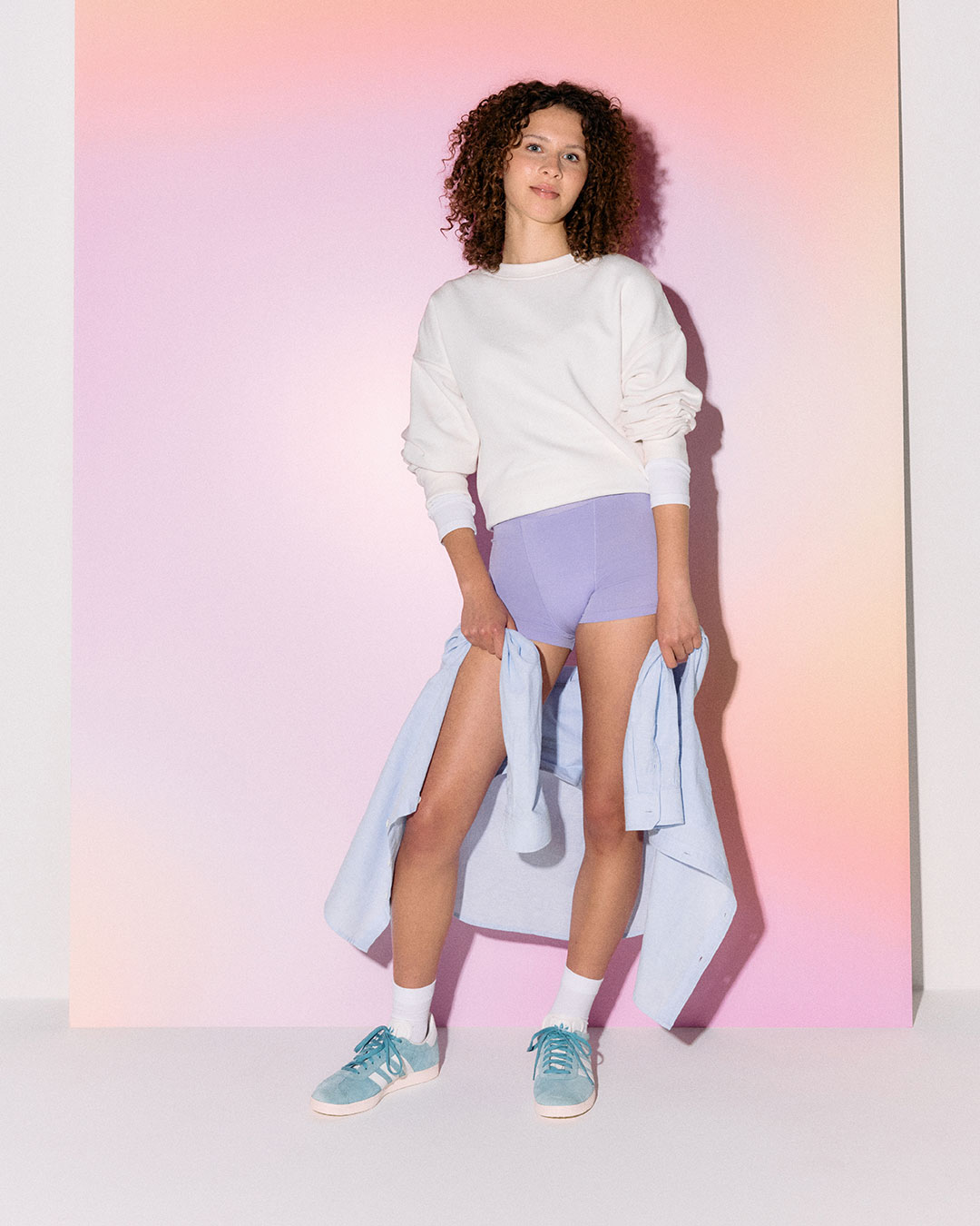 En person står framför en färgglad bakgrund med en ljus tröja och lila menstrosor.